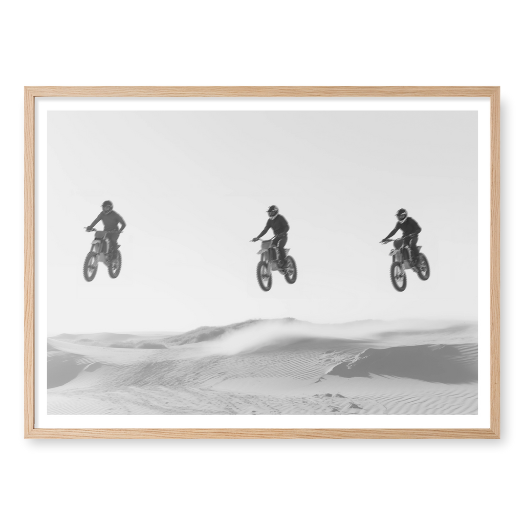 Three motocross riders jumping in a row over sand dunes, black and white art print titled Amigos in natural oak frame