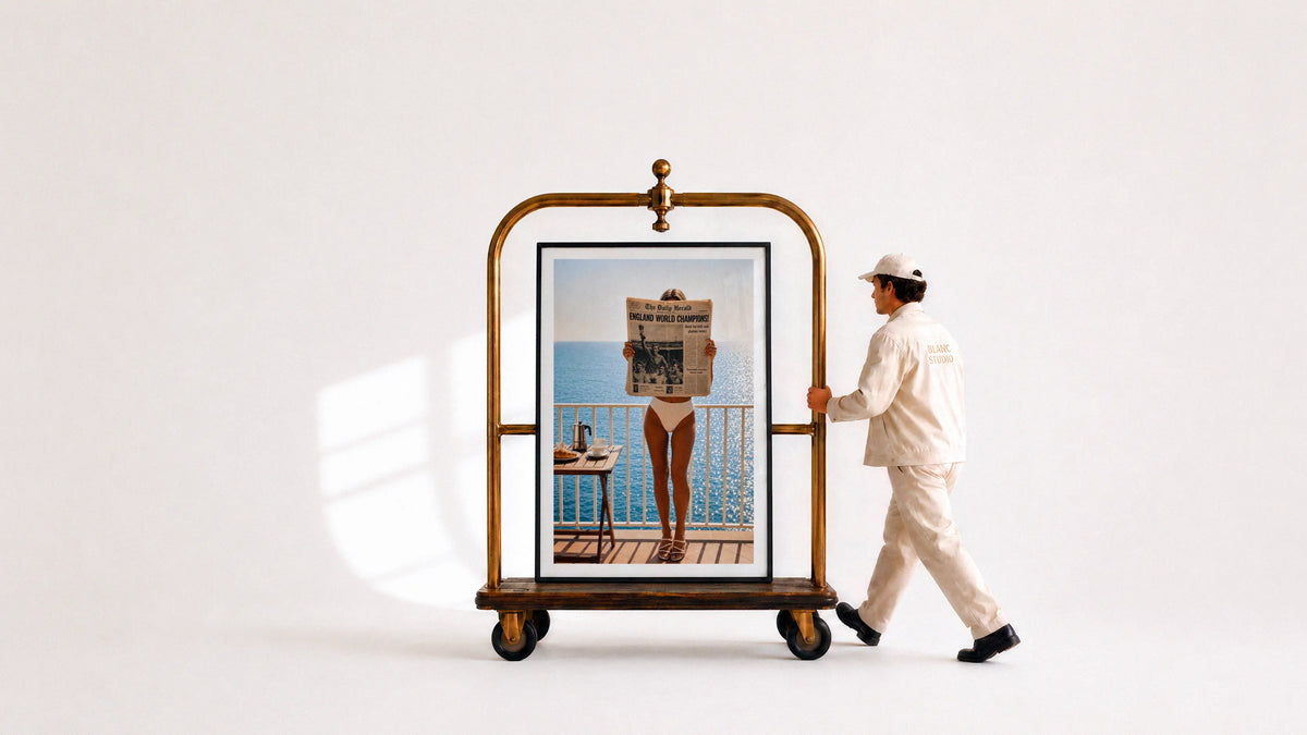Large black framed wall art print displayed on a vintage brass luggage trolley, pushed by a person wearing a Blanc Studio uniform