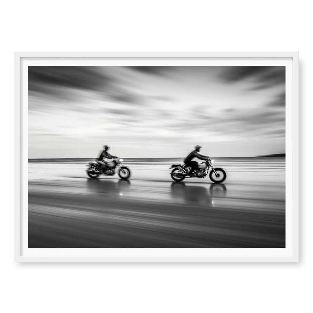 Panning shot of two riders on motorcycles racing along a wet beach with motion blur effect, black and white art print titled Partners in white frame