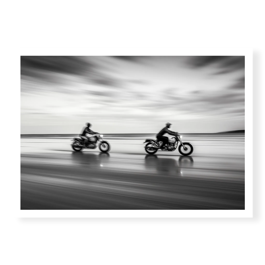 Panning shot of two riders on motorcycles racing along a wet beach with motion blur effect, black and white art print titled Partners